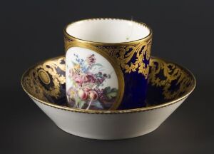  An ornate porcelain cup and saucer, with a white interior and a cobalt blue exterior embellished with gold patterns and an intricate floral design in a cartouche.