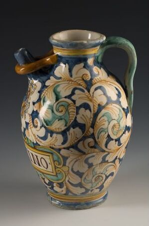  An ornate ceramic jug with a bulbous body decorated with interlocking green and blue patterns on an off-white background, featuring a green handle with an amber accent, set against a neutral backdrop.