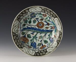  A decorative ceramic plate featuring a detailed pattern with blue, green, brown, and orange elements on a white background, exhibiting traditional floral and foliate motifs with a central undulating ribbon-like line.