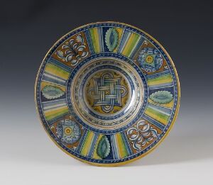  An intricately designed ceramic plate with a central emblem surrounded by concentric bands of geometric and organic patterns in vibrant blues, greens, yellows, and golds, displayed against a neutral background.