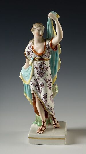  A porcelain figurine of a classical female figure in a flowing, patterned dress with a raised left arm, secured by a gold belt, standing on a small base, suggestive of Greek or Roman art influences.