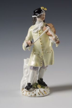  A delicate porcelain figurine of an 18th-century gentleman playing the violin, wearing a pale yellow coat with metallic button details and a black tricorn hat, against a neutral gray background.