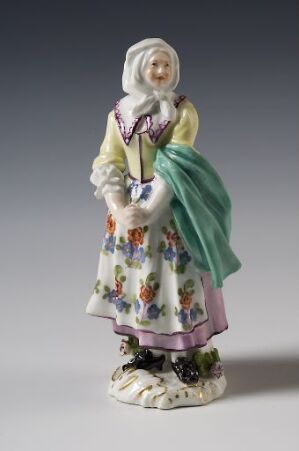  A porcelain figurine of an elderly woman in traditional European peasant attire with a white bonnet, floral dress, violet bodice, and green shawl, standing on a base adorned with flowers. Artist