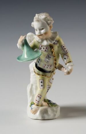  A small porcelain figurine of an 18th-century male character, in a decorative jacket and breeches with gold accents, holding an upturned green hat, and set against a light grey background. Artist name and title are unknown.