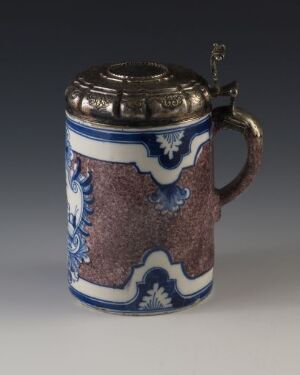  A traditional beer stein with a pewter lid and thumb lever. The body of the stein is decorated with intricate, curved dark blue and white patterns on the top and bottom, surrounding a mottled brown textured middle section suggestive of leather. The stein melds craft and functionality, embodying a sense of German tradition in its design.