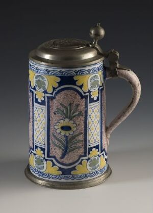 
 A traditional ceramic beer stein with a metal lid, featuring blue panels with purple and brown floral designs, a yellow and blue geometric border, and a muted brown handle, all against a white background.