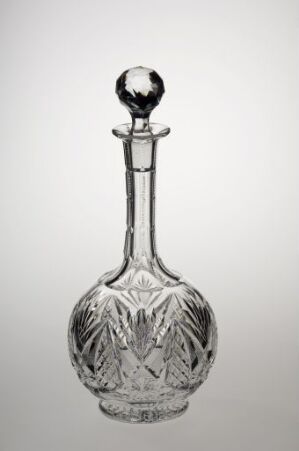  A clear, intricately cut glass decanter with a stopper, featuring leaf-like patterns on its bulbous body, placed against a light grey background.