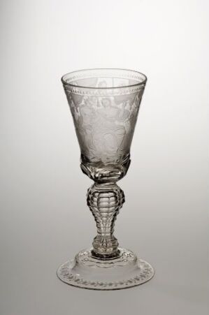  An ornate, clear glass goblet with a conical bowl featuring etched designs, an intricate bulbous stem, and a flared decorative base, set against a muted greyish-white background.