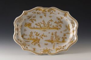  An antique porcelain serving dish with scalloped edges, detailed with pastoral gold scenes of leisure and stylized floral motifs on a creamy white background. The dish is displayed against a plain, dark background that accentuates the ornateness of the item.