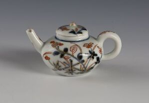  A small, white ceramic teapot adorned with intricate brown, orange, and black floral patterns, featuring a rounded body, handle, and a decorated lid, displayed against a soft grey background.