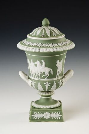  A jade-green classical urn made of ceramic or porcelain with white embossed figures and a matching lid with a decorative finial, flanked by two handles, standing on a square base with leaf-like motifs.