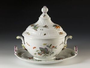  An elegantly shaped white porcelain soup tureen with a dome-shaped lid and ornamental finial, embellished with a naturalistic motif of insects and florals in soft oranges, greens, blues, and browns, presented on a matching oval tray against a neutral backdrop.