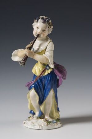  Porcelain figurine of a young boy in 18th-century attire playing a flute, with carefully detailed cream, yellow, purple, and cobalt blue clothing, and decorative floral patterns on a neutral gray background.