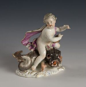  A porcelain figurine featuring a cherubic child riding a mythical creature with the body of a fish and the head of a dog, with a pink sash draped over the shoulder and detailed in soft pastel tones on a nuanced gray background. Artist and title are unknown.