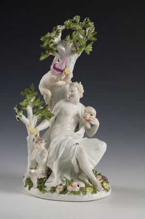  Delicate porcelain figurine depicting a classical male figure and child with green leaves, pastel flowers, and a goat with a purple ribbon.