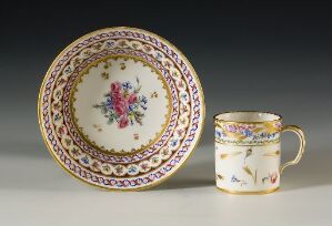  An ornate porcelain plate and cup set, featuring a central floral design with pinks, purples, and greens, accented by decorative gold and colored geometric bands. The cup matches the plate’s theme with similar colors and floral motifs.