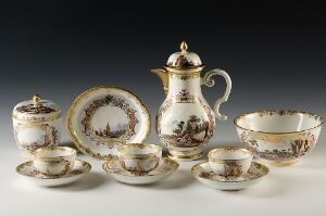  An antique porcelain tea set with sepia pastoral scenes and gold gilding on a light grey background. The set includes a cup with a saucer, a sugar bowl with a lid, a large teapot, a creamer, and a slop bowl.