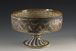  An ornate decorative bowl with earth tones and blue accents, featuring geometric and possibly figurative motifs, sitting on a flared pedestal base.