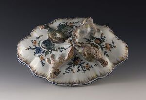  An ornate ceramic dish with scalloped edges featuring a raised central relief surrounded by dark floral motifs against a gradated off-white to pale cream background, accented with metallic gold or bronze edging.