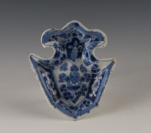  A blue and white ceramic piece with a unique, wavy-edged crest shape, intricately painted with cobalt blue floral designs against a white background, showcased against a neutral grey setting. Artist name and title are unknown.