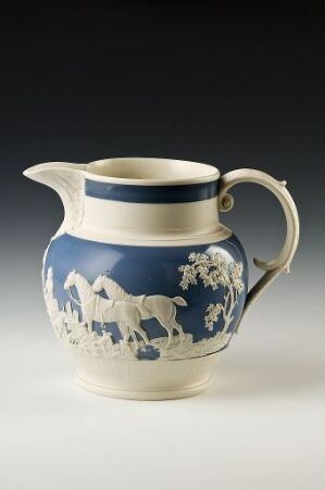  A ceramic pitcher with a deep cobalt blue and white color scheme, featuring classical white motifs on a blue band across the middle, set against a neutral grey gradient background.