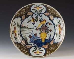  A vintage ceramic plate with central cobalt blue floral design, surrounded by golden yellow flowers, green leaves, and brown branches, with two yellow birds in flight, against an off-white background.