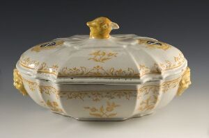  An oval-shaped decorative box made of white or cream-colored porcelain or ceramic, with gold floral or vine-like motifs and a translucent strip in the middle, featuring a gold or mustard yellow knob on the lid, against a neutral gray background. Artistname and title are unknown.