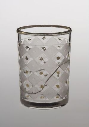  A clear glass cup with a gold-rimmed top and a decorative pattern of intersecting diamonds with small gold and white floral emblems at each junction, set against a soft grey background.