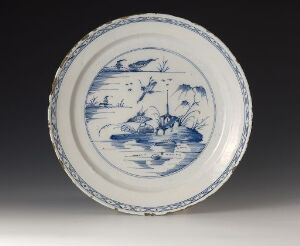  A round ceramic plate with a blue-and-white Eastern landscape design, featuring a boat with figures on a body of water, surrounded by a decorated rim with ornate cobalt blue motifs on a neutral grey backdrop.