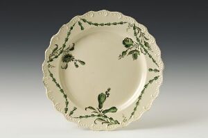  An ornate off-white ceramic plate with a scalloped edge, decorated with a symmetrical dark olive green pattern of foliage and small buds or berries, presented against a neutral grey background.