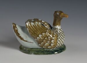  A ceramic figurine of a duck with detailed feathers in shades of golden brown, white, and deep brown, resting with wings partially spread on a green grass-like base.