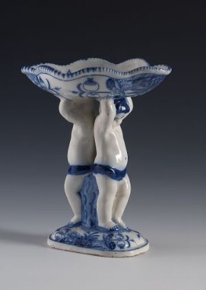  A ceramic figurine featuring two androgynous figures with a draped blue and white pattern at their hips supporting an ornate scalloped-edge bowl with a blue pattern on white, perhaps reminiscent of porcelain styles like Delftware.