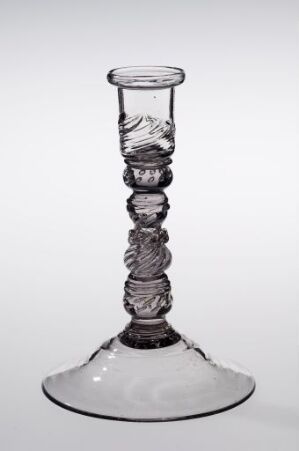  A clear glass candlestick holder with a broad base and a decorative stem featuring stacked spherical elements with etched designs, set against a soft, neutral background.