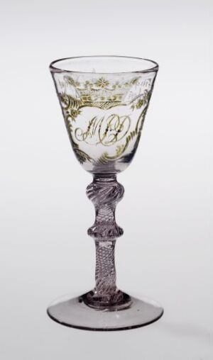  An intricate glass goblet with a decorative twisted stem and a detailed monogram surrounded by a leafy design etched into the bowl, against a plain light grey background.