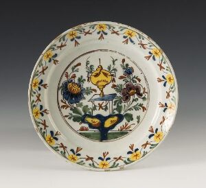  An intricately decorated vintage ceramic plate featuring a central floral design sprouting from a blue and yellow vase, with two colorful birds on either side amidst a repeating floral motif around the rim.