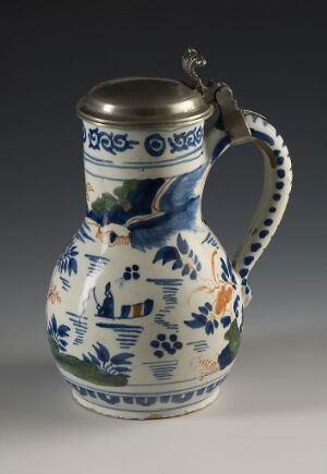  A ceramic jug with pewter lid, featuring a blue, orange, and green floral motif, with blue-painted houses and birds, against a neutral gray background.