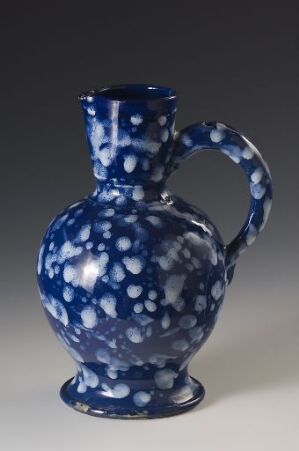  A glossy, cobalt blue ceramic jug with small, irregular white spots covering its surface stands against a neutral gray background. The jug has an elegant, curved handle, a bulbous base, and a flaring mouth.