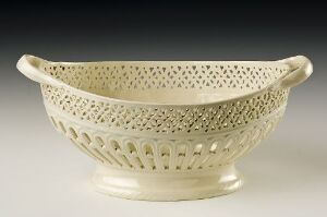  An intricately designed, footed, oval bowl with a lace-like, perforated pattern, finished in a light cream color, against a neutral gray background.