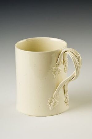  An off-white cylindrical ceramic mug with an ornate 'S'-curved handle and subtle floral embossments on the body, set against a light background.