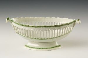  An oval-shaped cream-colored ceramic bowl with a delicate green trim on the rim and base, vertical piercings creating an openwork design along the sides, scalloped edges, and two matching green handles.