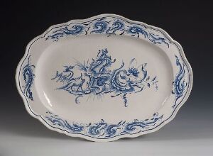  An oval-shaped white-glazed faience serving platter with hand-painted cobalt blue floral and scroll decorations around the edges and in the center, showcased against a neutral background. The artist's name is unknown.