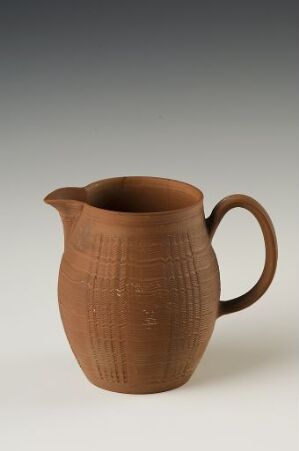  A rustic brown ceramic pitcher with a textured surface made of fine vertical lines, a curved spout, and a single handle, set against a neutral gray background. The title and artist name are unknown.