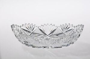  A clear crystal bowl with scalloped edges and intricate cut glass designs sits on a light grey background. The bowl is oval-shaped and radiates with its reflective cuts and elegant details.