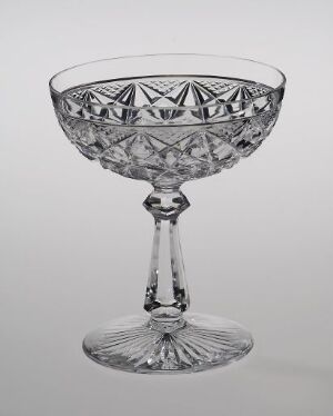  A crystal dessert glass with intricate cut-glass patterns, created by Hadeland Glassverk, stands with a faceted bowl, a slender stem with a spherical detail, and a patterned base, reflecting light and showcasing the artistry of blown and ground glass technique.