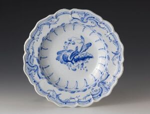  A white and blue faience plate with a scalloped edge, featuring a stylized blue floral motif at the center, surrounded by an intricate lattice-patterned design in cobalt blue. The item is known as "Det rehnska mönsteret."