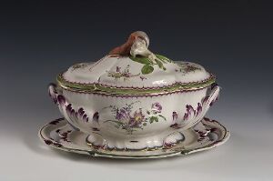  An antique porcelain tureen with floral motifs in purple, pink, and green, with gold trim, displayed on an oval saucer, all against a neutral gray background.