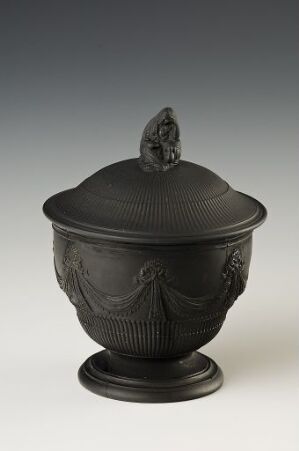  A dark grey or black neoclassical urn with relief decorations and a pedestal base, featuring a ribbed lid topped with a small sculptural finial. The urn exhibits elegance and a traditional classical design, possibly made of metal or ceramic. Artist name and title are unknown.