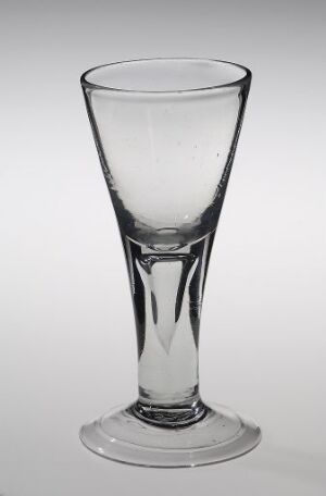  An empty, clear glass with a flared rim and a tapered shape standing against a light gray background, demonstrating its transparent quality and reflecting the surrounding light.