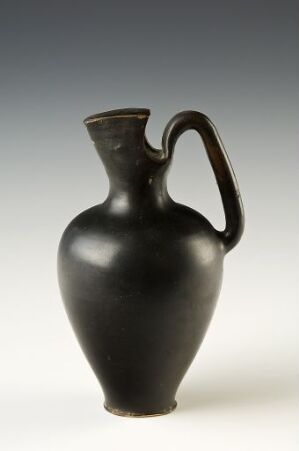  A simple black ceramic pitcher with a smooth finish and a single handle against a gradient gray background, representing an object with historical or ancient design elements.