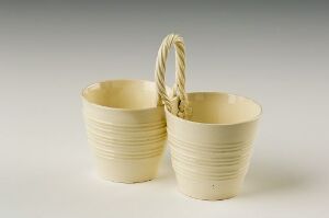  Two light creamy ceramic cups connected by a single twisted handle with horizontal lines wrapping around the cups, set against an off-white background. Artist name and title are unknown.